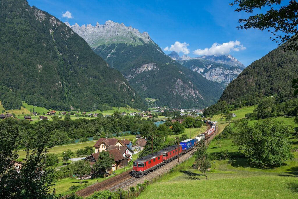 ขบวนรถไฟบรรทุกสินค้าสีแดงบนเส้นทาง Gotthard ใกล้เมือง Erstfeld ท่ามกลางหุบเขาสวิตเซอร์แลนด์
