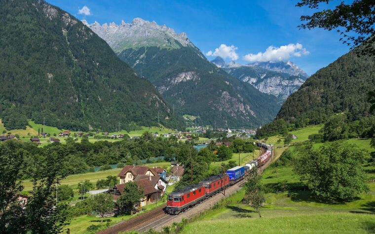 ขบวนรถไฟบรรทุกสินค้าสีแดงบนเส้นทาง Gotthard ใกล้เมือง Erstfeld ท่ามกลางหุบเขาสวิตเซอร์แลนด์